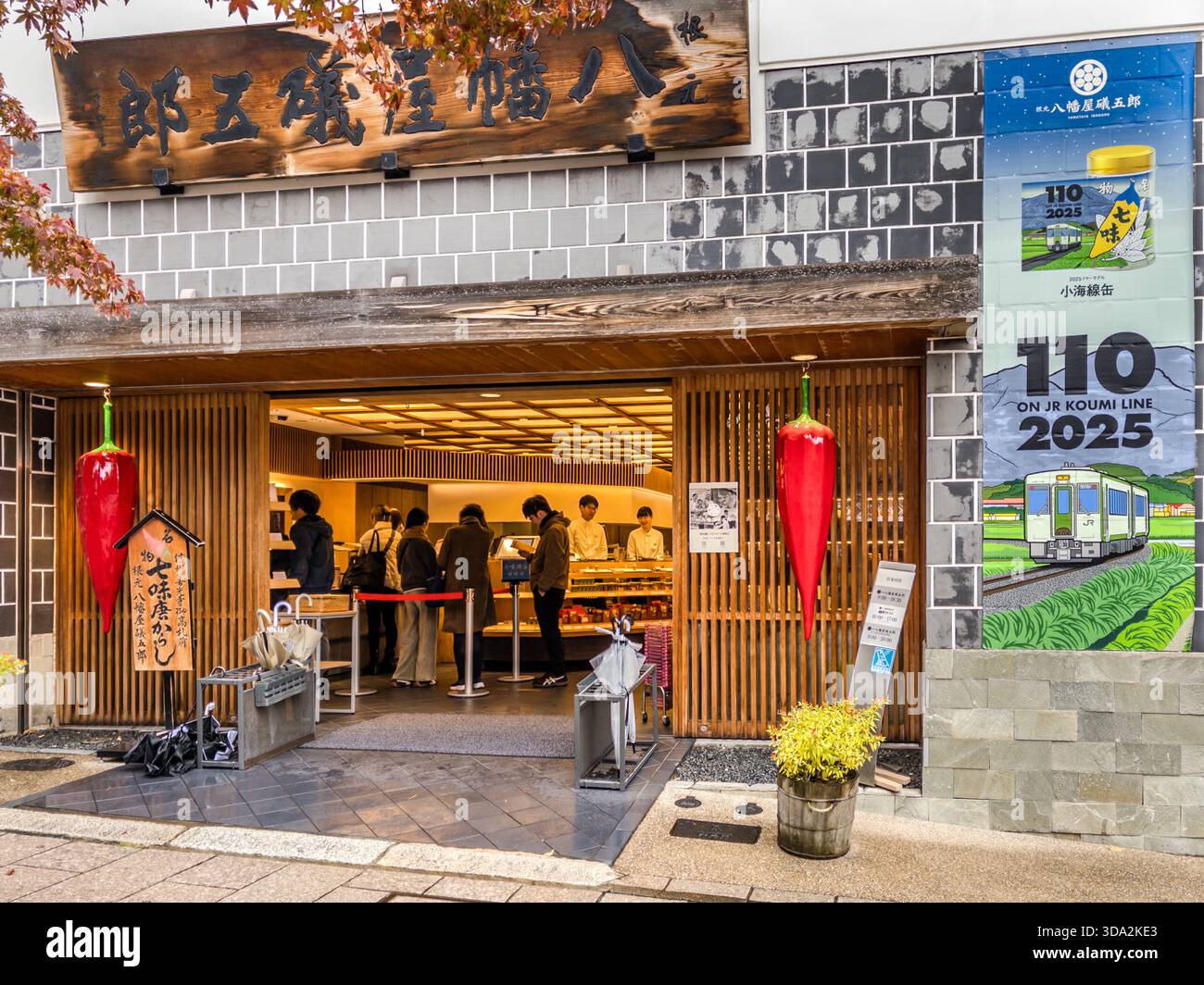 Two oversized red chilli peppers flank the wooden entrance of the Yawataya Isogoro (八幡屋礒五郎) flagship store in Daimon-cho, five minutes' walk from Zenkoji Temple (善光寺). The shop was founded in 1736 by the hemp and washi paper merchant Kan'emon Muroga, who later discovered shichimi togarashi in Edo. A banner beside the entrance advertises the JR Koumi Line's 110th anniversary in 2025, its train livery decorated with the store's spice tin design. Stock Photo