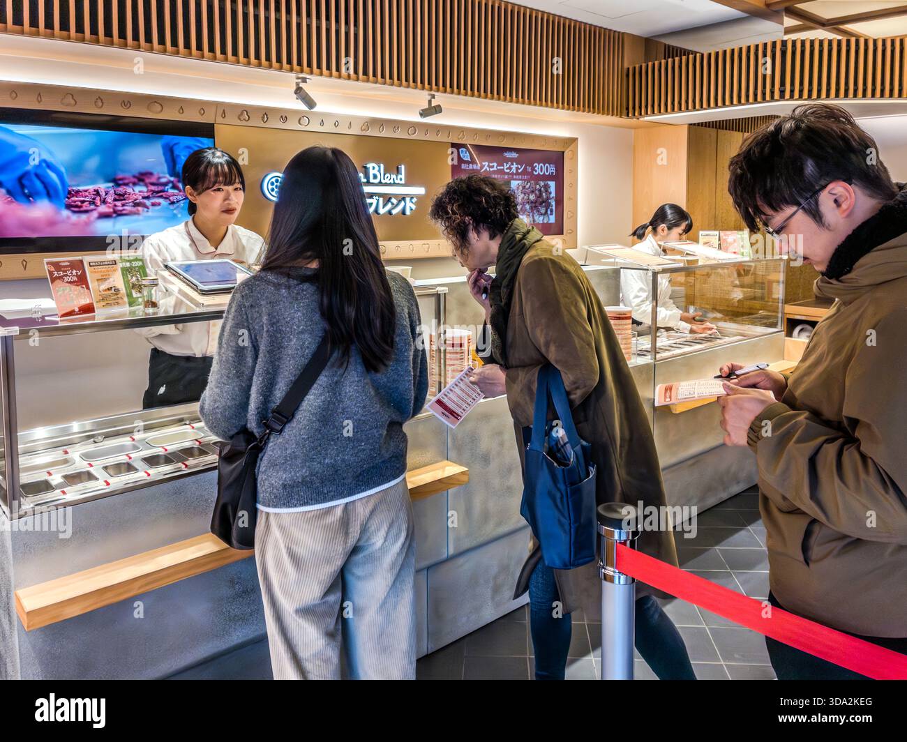 Customers queue behind a red rope at the counter of the Yawataya Isogoro (八幡屋礒五郎) flagship store on Daimon-cho in Nagano City, where staff serve the shop's shichimi spice blends. Stock Photo