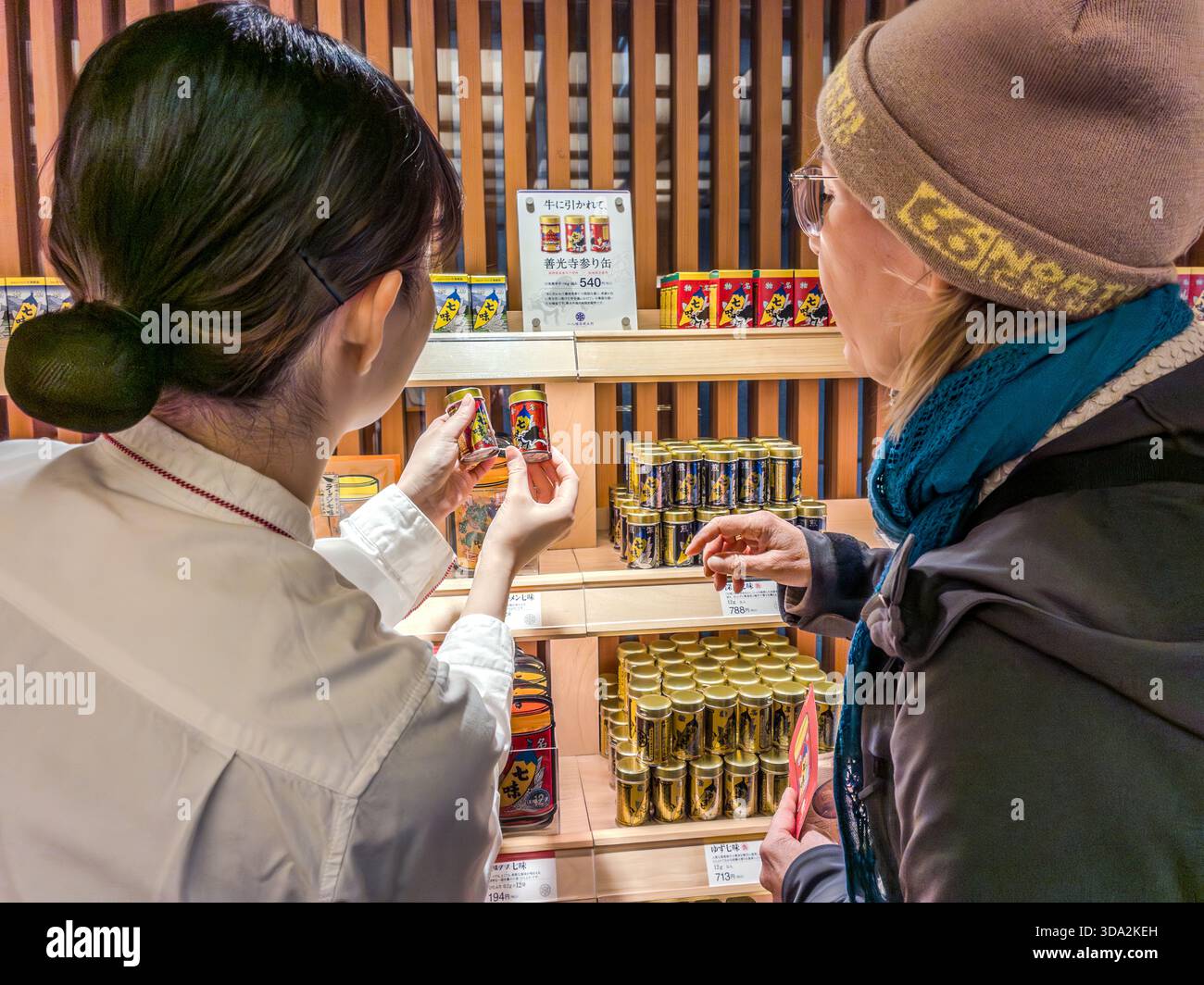 A shop assistant helps a customer choose a shichimi spice tin at the Yawataya Isogoro (八幡屋礒五郎) flagship store on Daimon-cho in Nagano City. The red and gold tin, decorated with a large chilli motif and an image of Zenkoji Temple (善光寺) on the back, has kept the same design for decades. Stock Photo