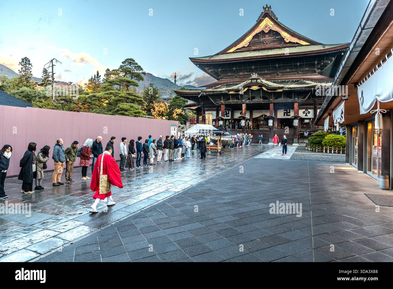 Pilgrims queue in front of Zenkōji Temple, one of Japan's most important Buddhist temples and the historic heart of Nagano, Nagano Prefecture, Japan. A monk in a red robe crosses the courtyard. Founded in the 7th century, Zenkōji is regarded as one of the country's oldest and best-known temples; the city of Nagano grew up around it as a temple town. Stock Photo
