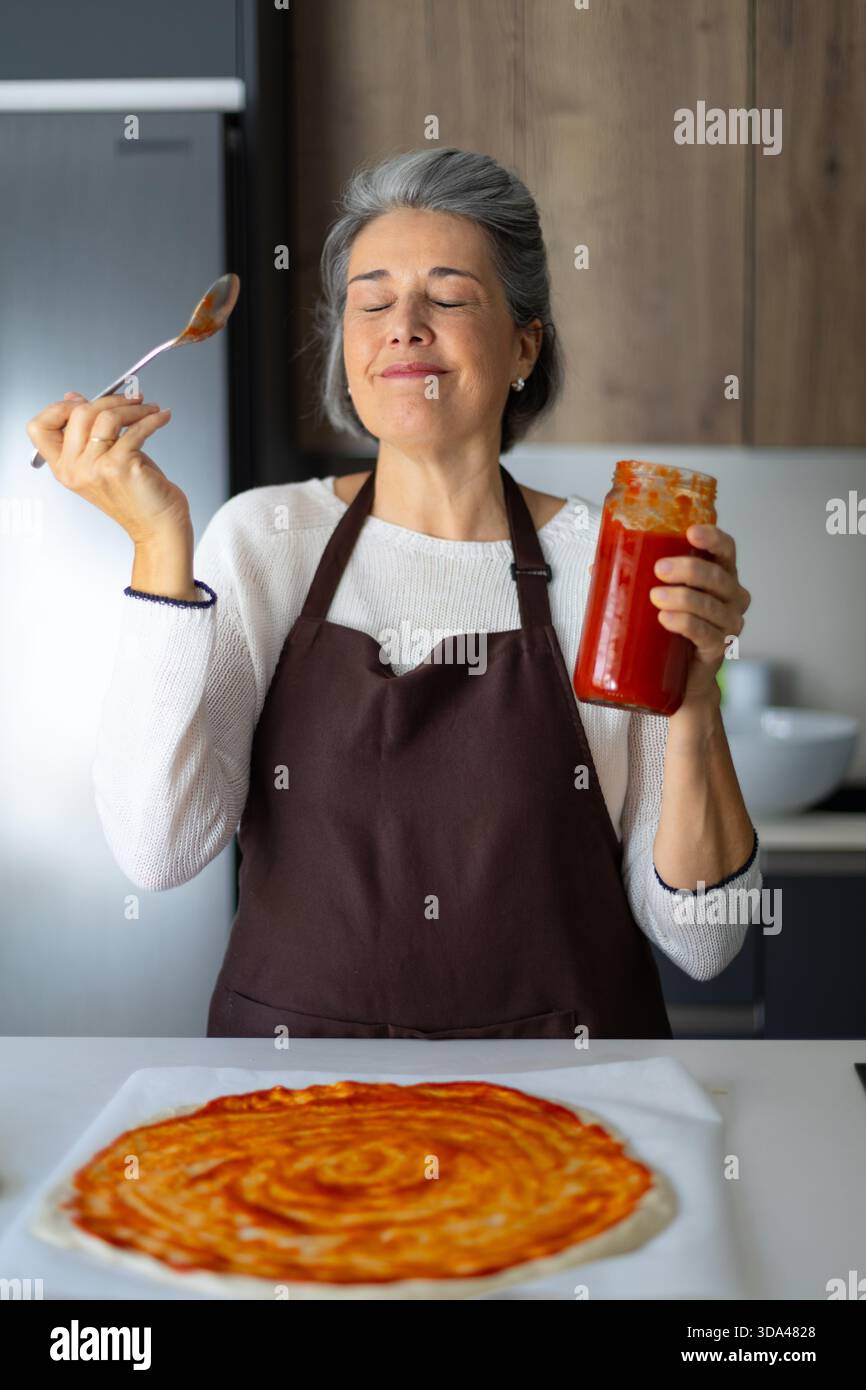 Woman in kitchen smelling delicious tomato sauce while preparing homemade pizza, representing passion for cooking and food Stock Photo