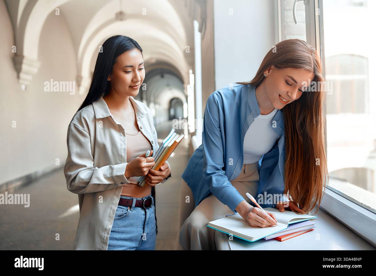 Two students collaborate on coursework, building a friendship in a university hallway, focusing on learning and teamwork. Their collaboration symboliz Stock Photo