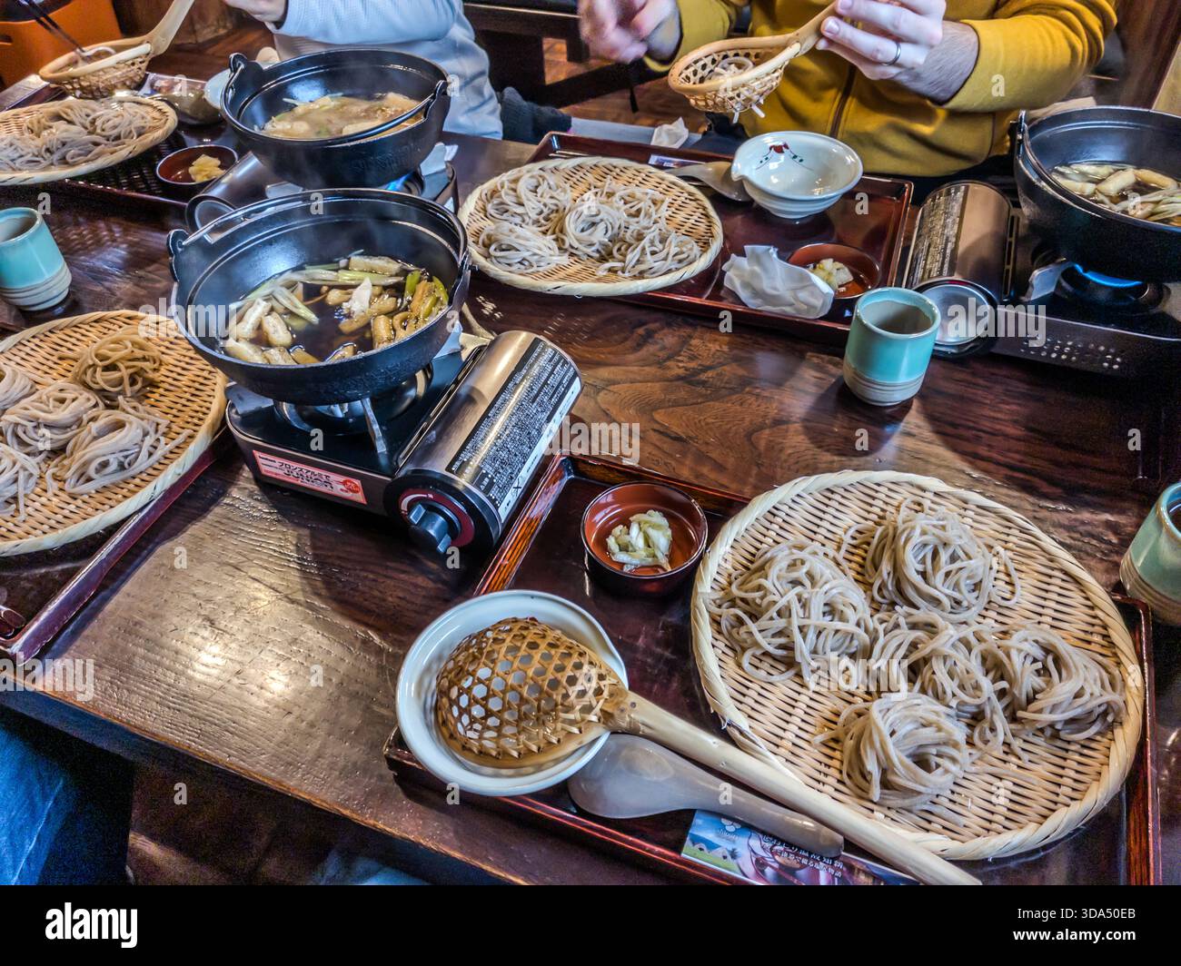 Bamboo trays of cold soba noodles cover a restaurant table in Shiojiri, Nagano Prefecture, next to small gas stoves that keep dashi dipping broth warm. Nagano is Japan's leading soba-producing prefecture; the cool mountain air and nutrient-rich soil of its highlands create ideal conditions for growing buckwheat, seasoned here with shichimi spice. Stock Photo