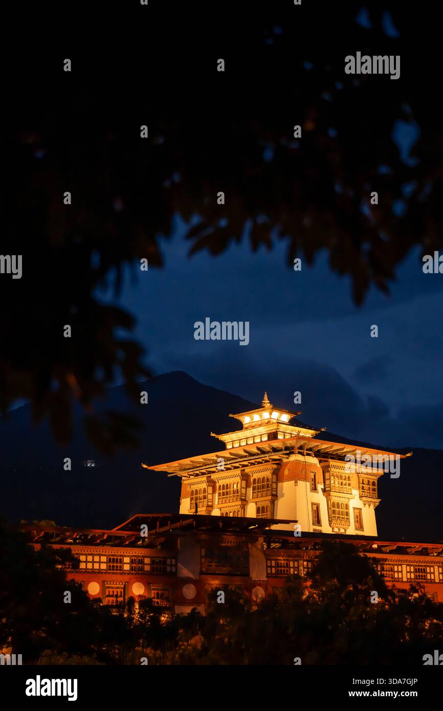 View of golden light illuminating the intricate architecture of a traditional building against the dark sky and silhouetted mountains, Punakha, Bhutan Stock Photo