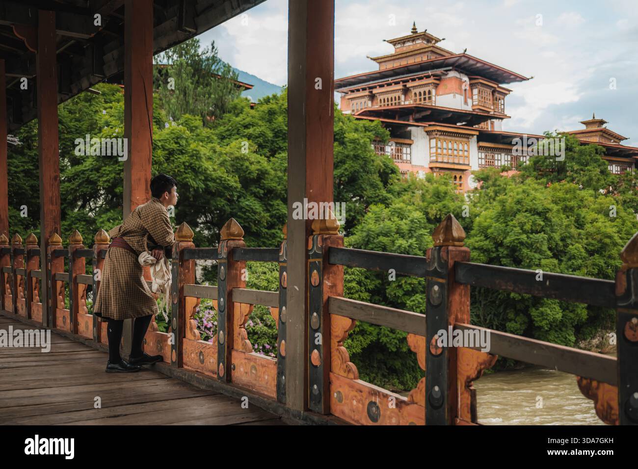 View of a person in traditional dress gazing at a multi-tiered building nestled among lush greenery from an ornate wooden bridge, Punakha, Bhutan. Stock Photo