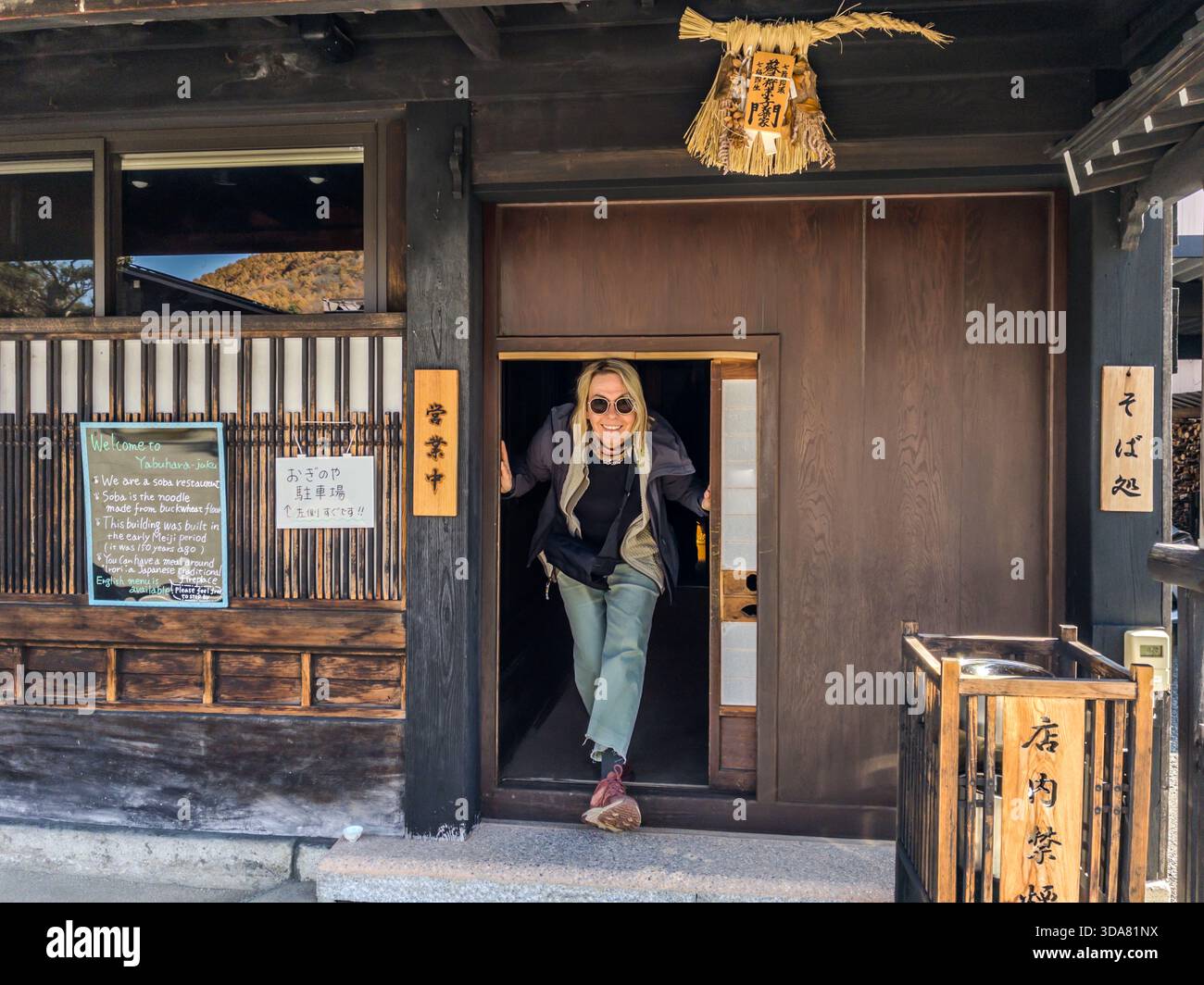 A traditional wooden house with a narrow entrance door holds a soba noodle restaurant on the old Nakasendo (中山道) in the Kiso Valley, Nagano Prefecture. A sign by the door dates the building to the early Meiji era, more than 100 years old. Shichimi spice and soba noodles have gone together since the Edo period: the seven ingredients built around chilli were once used as a remedy to warm the body and stimulate blood circulation. Angela Berg laughs as she steps out through the low doorway. Stock Photo