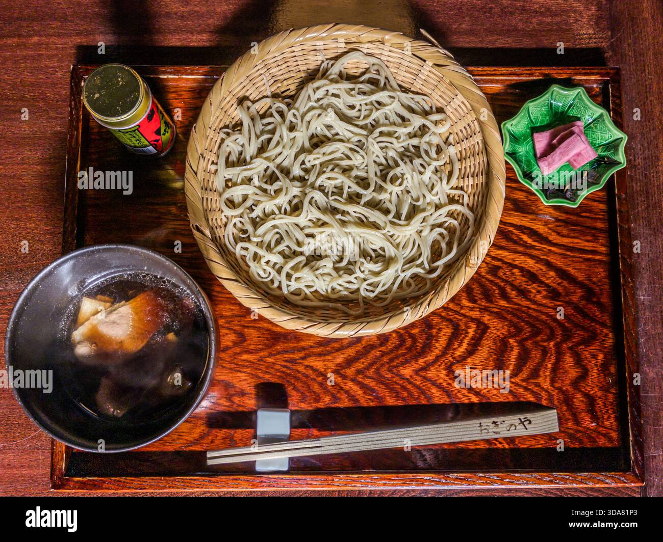 Cold soba noodles rest on a round bamboo tray, served with dashi broth for dipping and a red tin of shichimi spice from Yawataya Isogoro (八幡屋礒五郎). Nagano is Japan's leading soba-producing prefecture: the cool mountain air and nutrient-rich soil of its highlands create ideal conditions for growing buckwheat. Kiso, Nagano Prefecture, Japan. Stock Photo