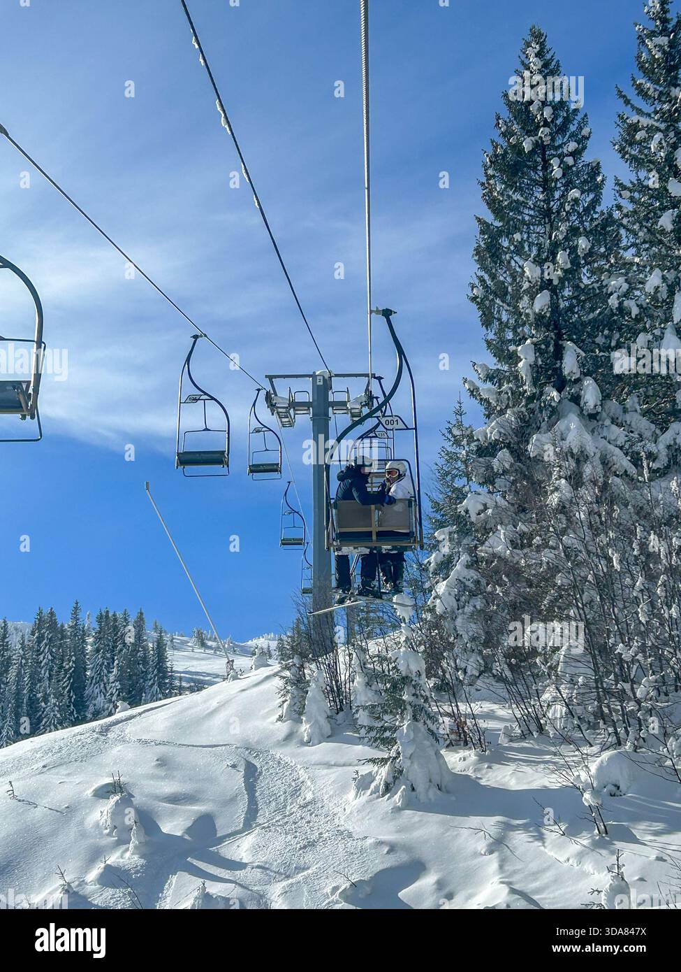 A couple enjoys a peaceful ride on the Ogorjelica double chairlift at Jahorina ski resort, surrounded by fresh snow, evergreen trees, and clear blue s - Smartphone Captured Stock Image