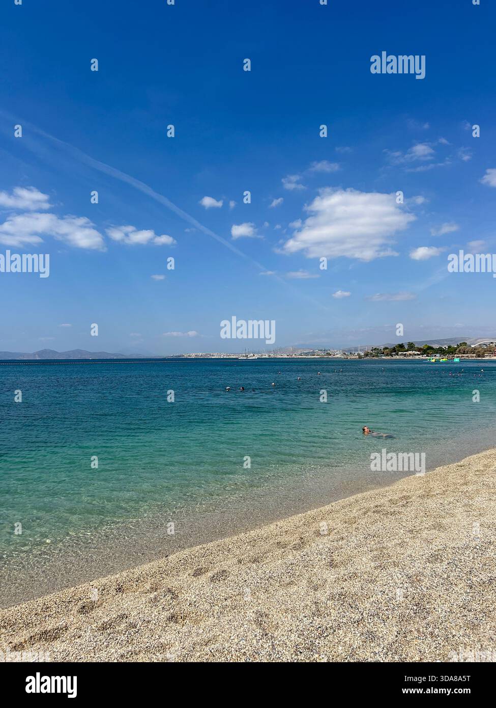 Sunny day at a pebble beach with crystal-clear turquoise water, calm waves, and swimmers enjoying the peaceful shoreline under a bright blue sky. - Smartphone Captured Stock Image