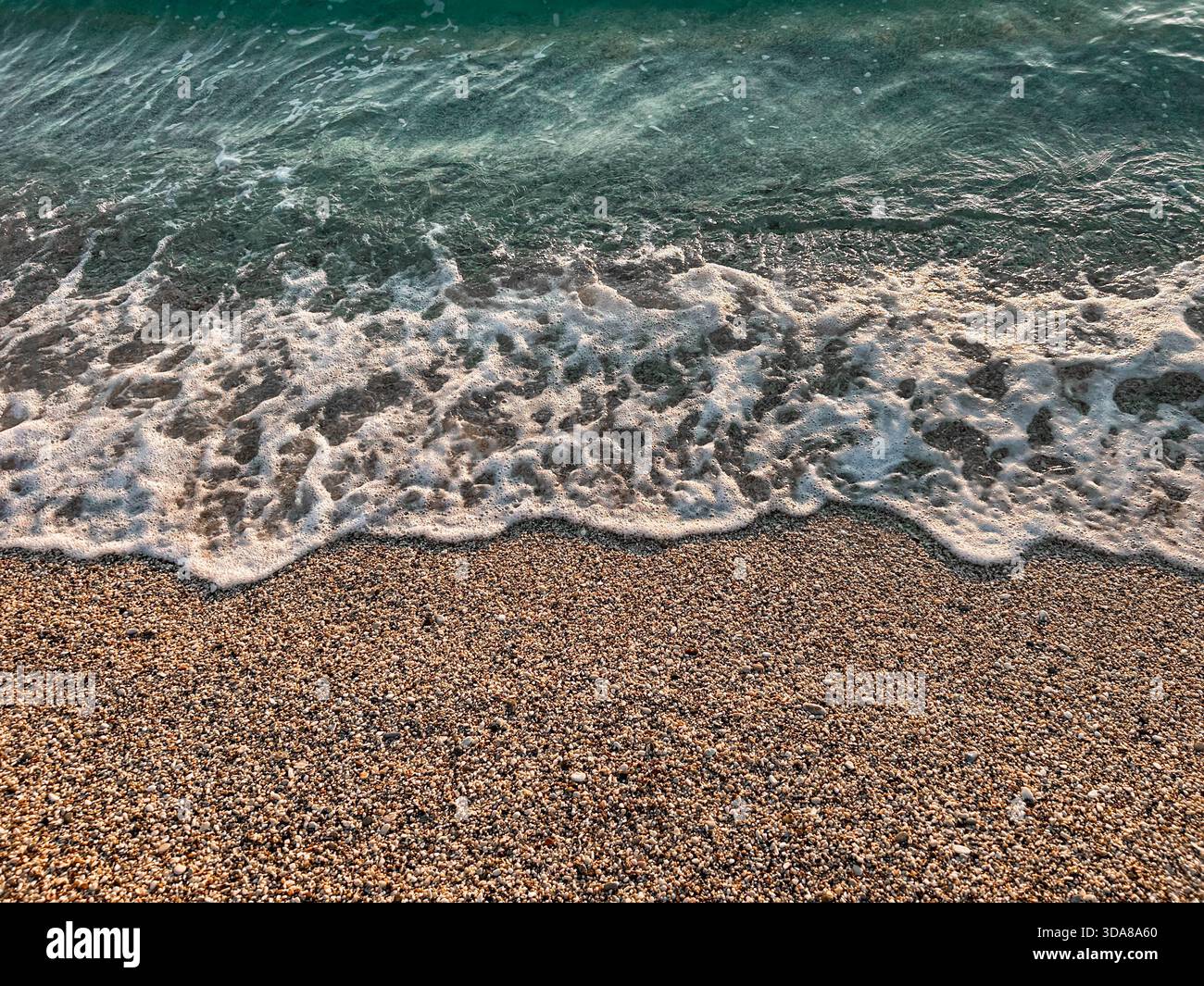 Shallow turquoise water meets a pebbled sandy beach as small waves retreat, revealing detailed textures and natural seaside colors. - Smartphone Captured Stock Image