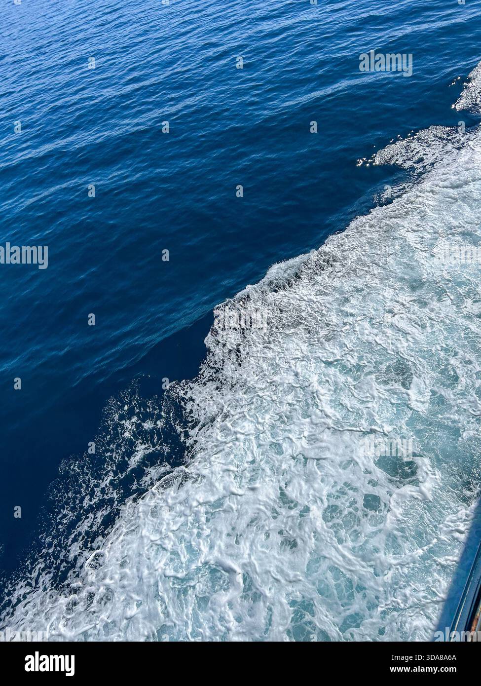 Foamy white waves trail across deep blue ocean water, captured from a moving boat on a bright, calm day. - Smartphone Captured Stock Image