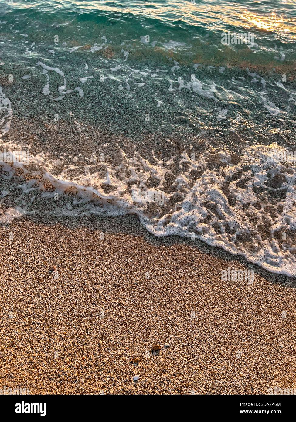 Soft wave foam washes over a coarse pebble shore, showcasing the transition from clear blue water to textured beach surface in warm natural light. - Smartphone Captured Stock Image
