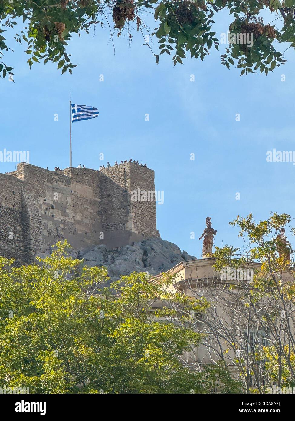 View of the Greek flag waving above the Acropolis, framed by trees, with a statue of goddess Athena in the foreground under a clear blue sky. - Smartphone Captured Stock Image