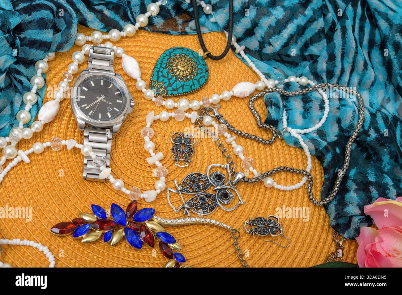 Assorted jewelry and wristwatch arranged on a woven straw mat with blue fabric and flowers. Stock Photo