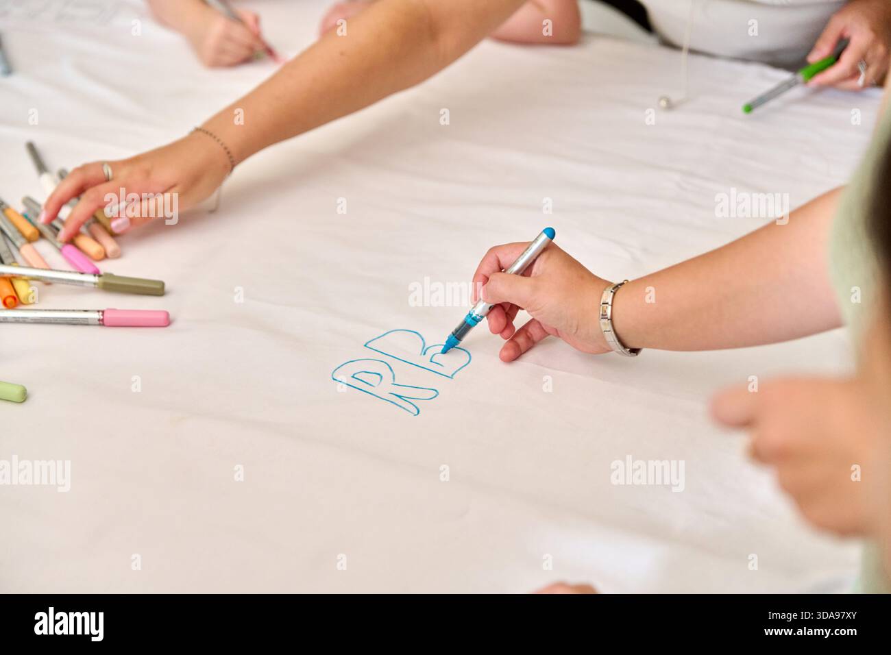 Multiple hands actively drawing and coloring on large white paper surface using various colored markers. Bright lighting highlights creative collabora Stock Photo