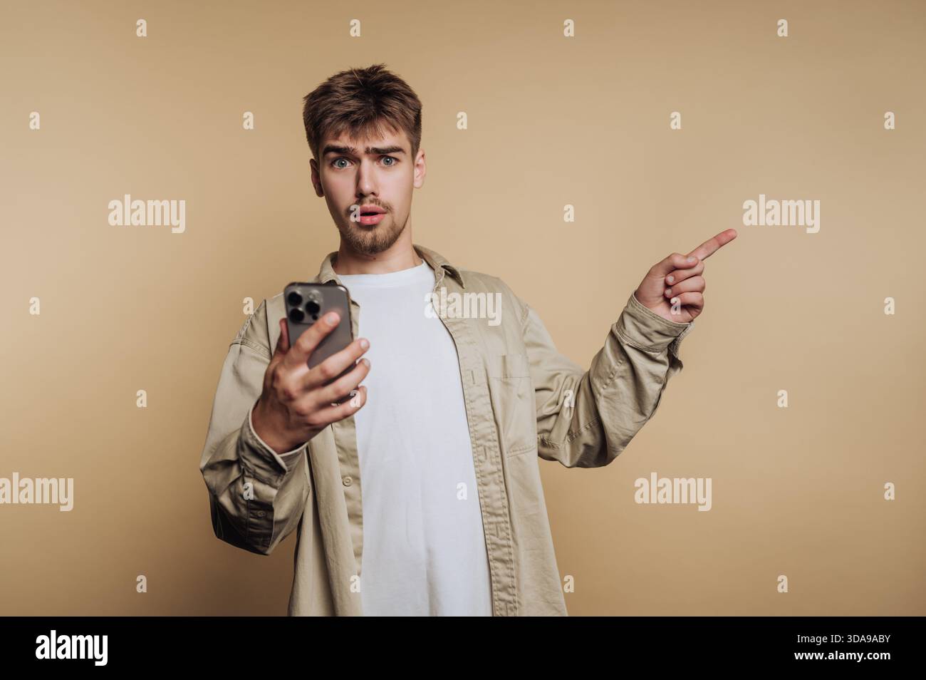 Young man with surprised expression holding smartphone and pointing. Stock Photo