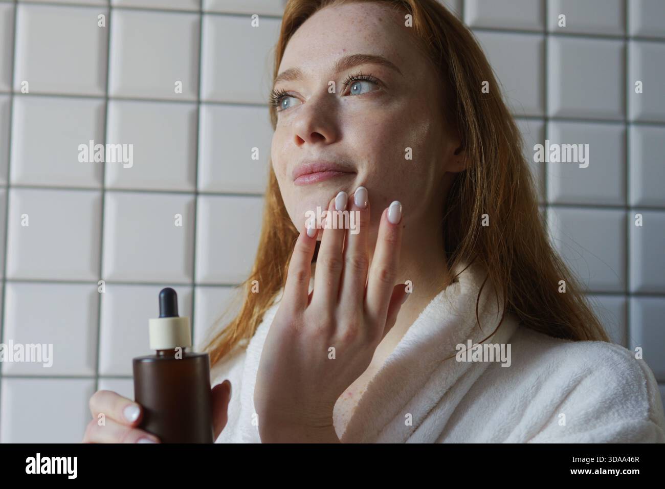 Woman In Robe Uses Dropper For Skincare, Relaxed Woman Applies Nourishing Lotion To Her Chin Carefully Stock Photo