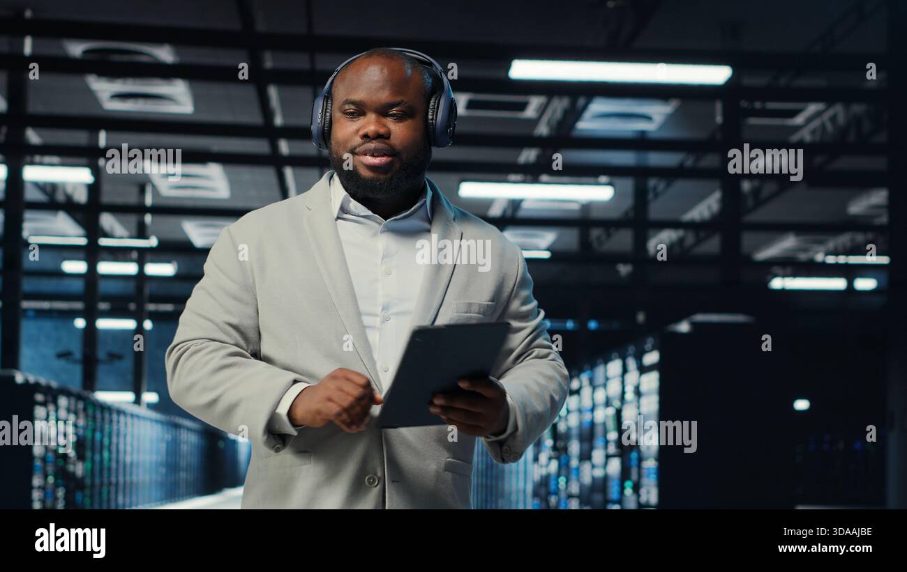 Data center admin wearing headphones using tablet to identify bottlenecks. Server farm IT expert listening music while troubleshooting bugs, implementing optimizations, camera B Stock Photo