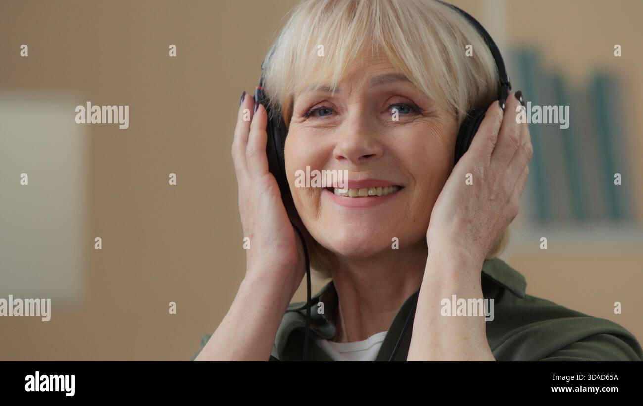Old Caucasian woman in headphones smiling listening music in headphones looking at camera senior female retired lady happy listen audio grandmother in Stock Photo