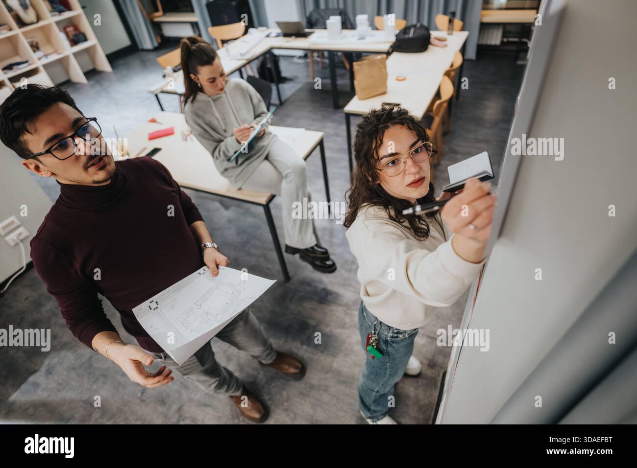 Students collaborate in a modern studio, reviewing blueprints and writing on a whiteboard Stock Photo