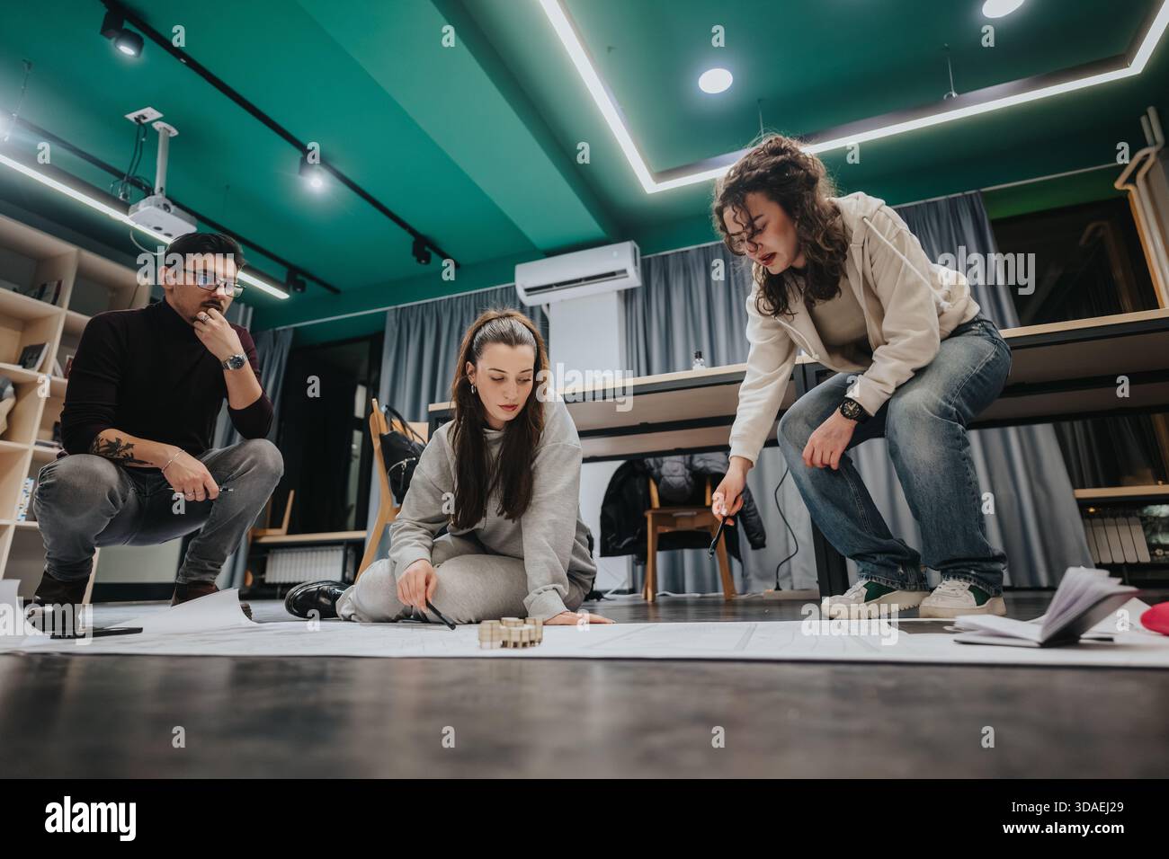 Three students collaborate in a modern studio, reviewing plans and building a model during a group study session Stock Photo