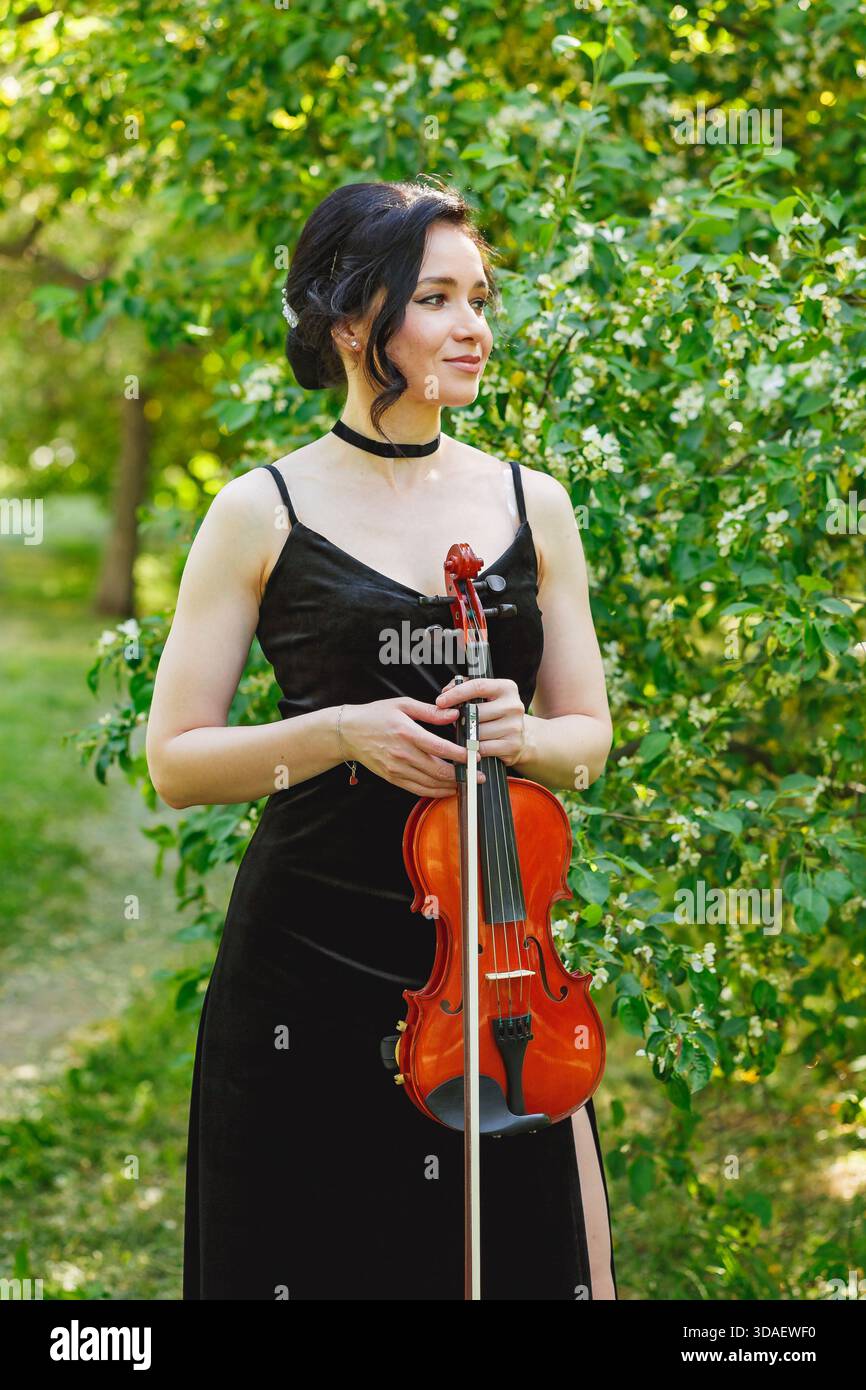 A violinist wearing a black dress poses elegantly with her violin in a lush garden setting, surrounded by vibrant green foliage and delicate white flo Stock Photo