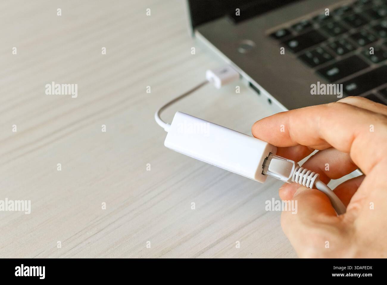 Close-up of a hand connecting a white Ethernet cable to a USB Ethernet adapter plugged into a silver laptop on a light desk, illustrating quick wired Stock Photo