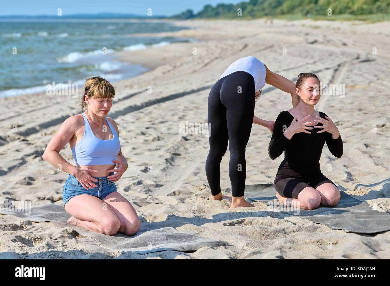 Two young women engage in pranayama breathing and body tapping exercises on sandy Baltic Sea shore under bright daylight. Calm water and green backgro Stock Photo