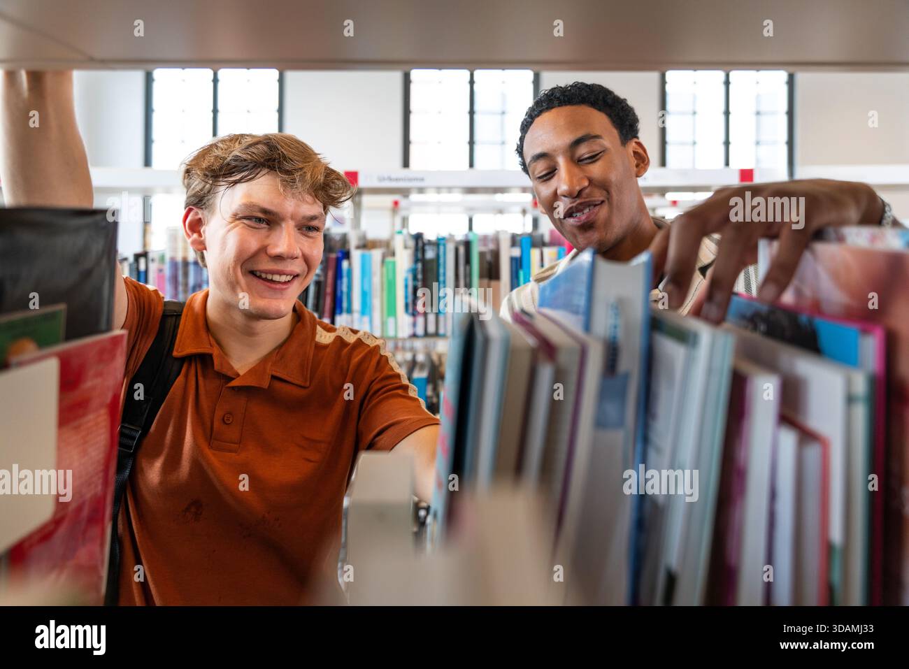 Two young diverse male students standing in a library, smiling and searching for books on shelves, representing education, knowledge, diversity, and a Stock Photo