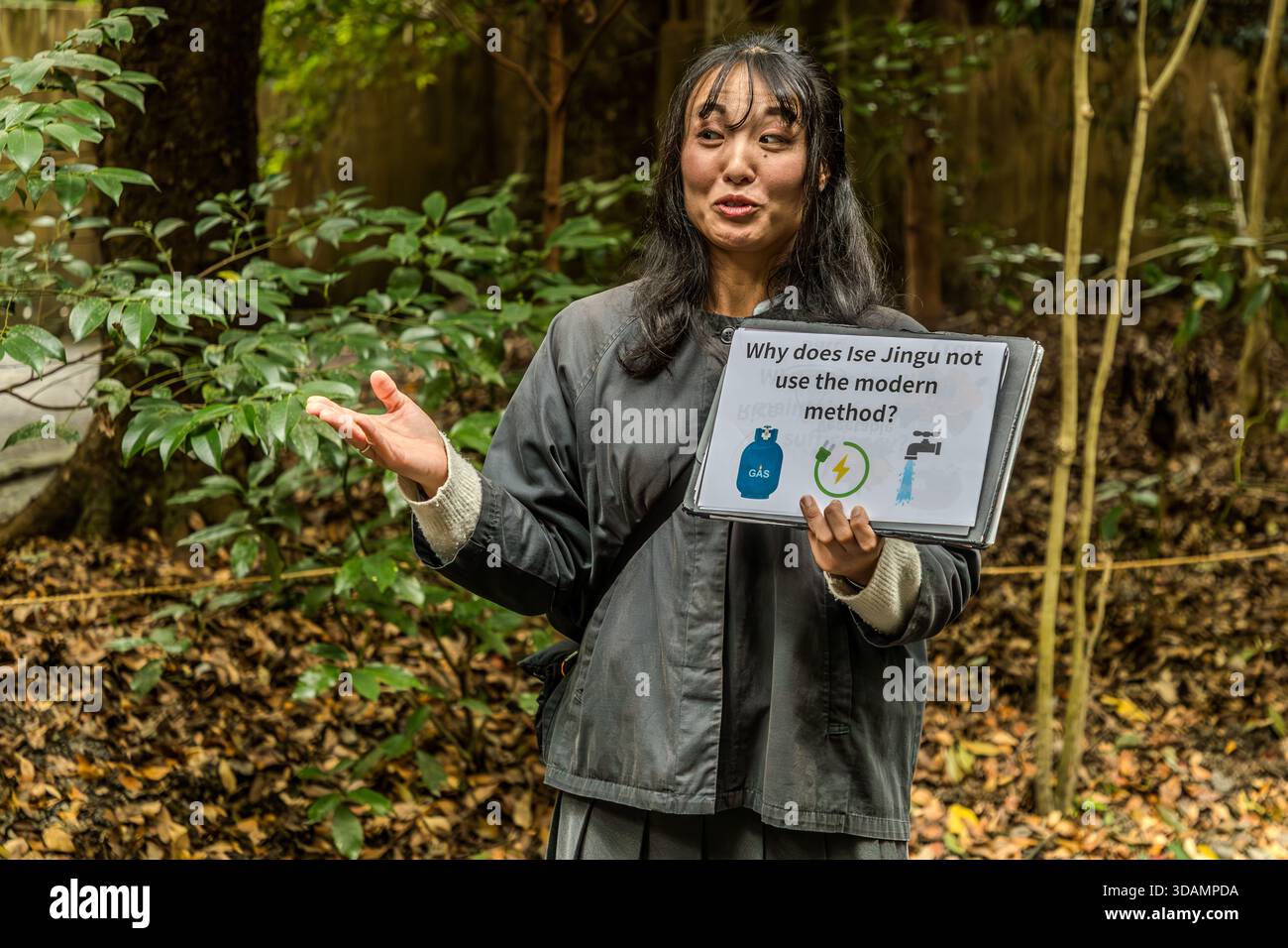 Tour guide Chifumi Takahashi holds up a sign asking why Ise Jingu does not use modern methods, illustrated with icons for gas, electricity and running water. Ise, Mie Prefecture, Japan. Stock Photo