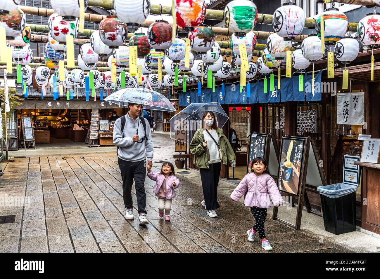 A family strolls through Oharai-machi, the historic pilgrimage and shopping street in front of the Naikū of Ise-jingū, Ise, Mie Prefecture, Japan. Hand-painted paper lanterns, chōchin, hang from bamboo poles above the alley. The traditional wooden shopfronts date back to the Edo period; for centuries the street has served as the access route for pilgrims, Okagemairi, to Japan's most important Shinto shrine. Stock Photo