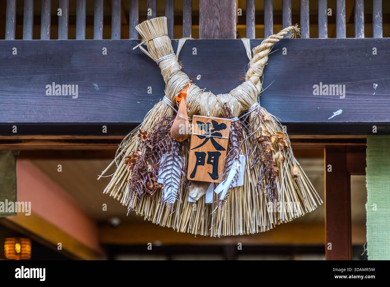 An Ise-Shimekazari with a wooden plaque inscribed "笑門" (Shōmon, "Gate of Laughter") hangs on a lintel in Oharai-machi, Ise, Mie Prefecture, Japan. The inscription alludes to the Japanese proverb "Warau kado ni wa fuku kitaru", "good fortune comes to the house from which laughter comes". The amulet, woven from rice straw with dried plants and strips of shide paper, is a form of the shimenawa purification symbol characteristic of the Ise region. The combination of Shinto purification symbolism and everyday wisdom is typical of religious practice in Japan. Stock Photo