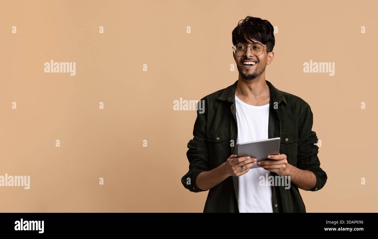 Young Man Smiles While Holding a Tablet Against a Simple Beige Background in a Bright Indoor Setting Stock Photo