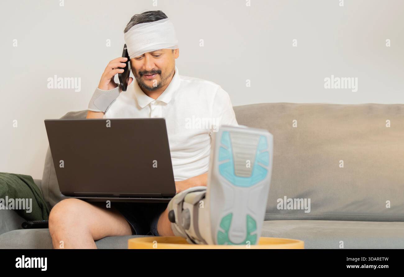 Positive man enjoying sick leave despite physical injury. Happy smiling young guy with broken leg and head sitting in sofa at home, using mobile phone Stock Photo