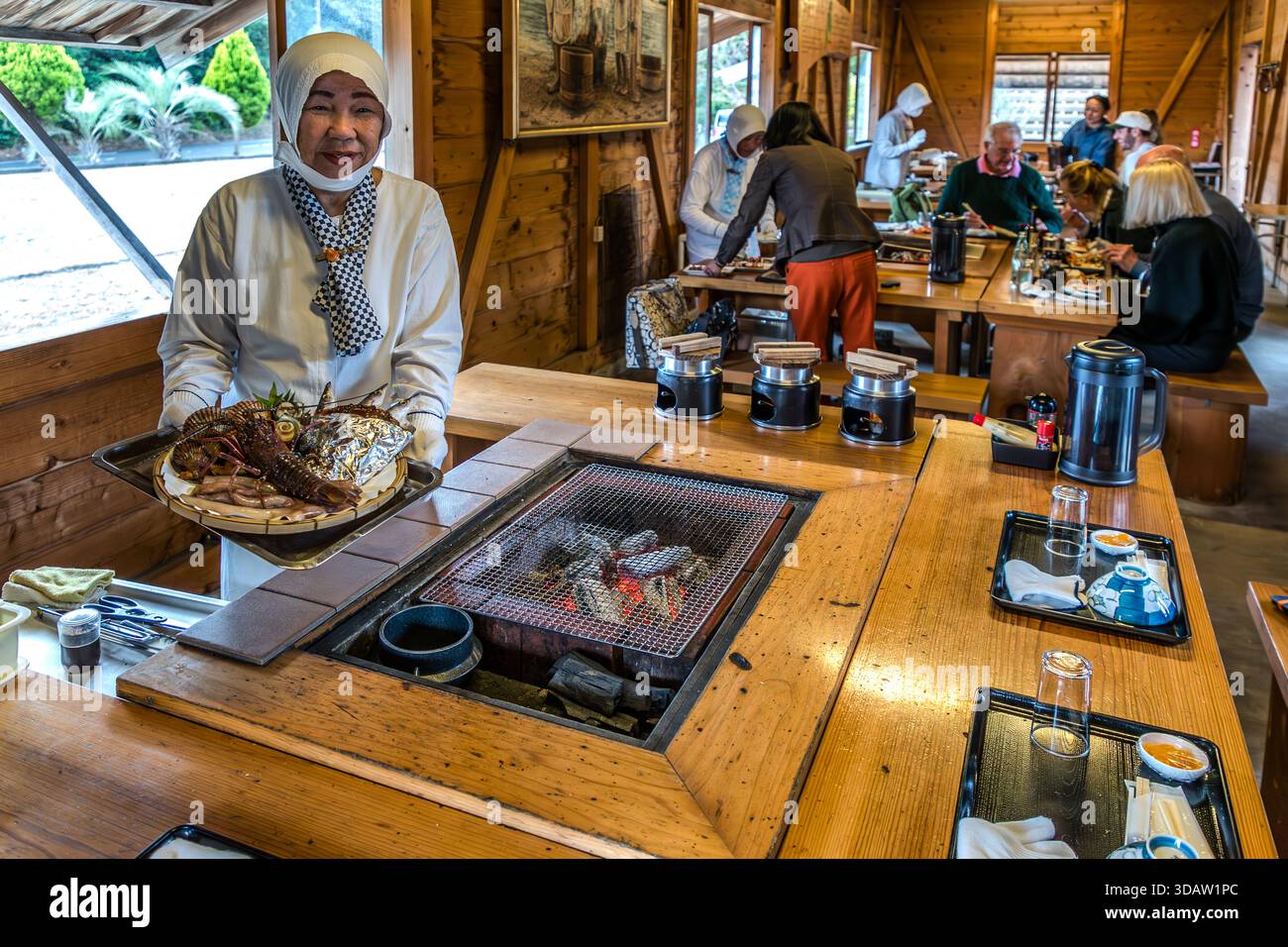 Ama diver Kimiyo Hayashi prepares fresh seafood on a charcoal stove right in front of the guests, in a replica of a traditional ama hut on the coast of Mie, Shima, Mie Prefecture, Japan. Hayashi, 71, has worked as an Ama diver for 56 years, having started at the age of 15. During the Ama Hut Experience, Ama divers grill freshly caught seafood for guests in a replica ama hut. Stock Photo
