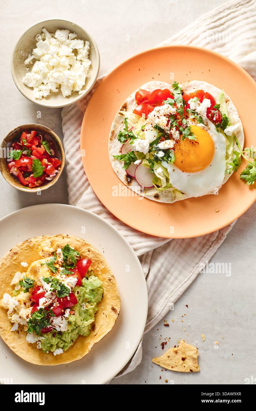 Top view of two plates with Rancheros eggs. One features a sunny-side-up egg on tortilla with fresh veggies, served on orange ceramic. The other has s Stock Photo