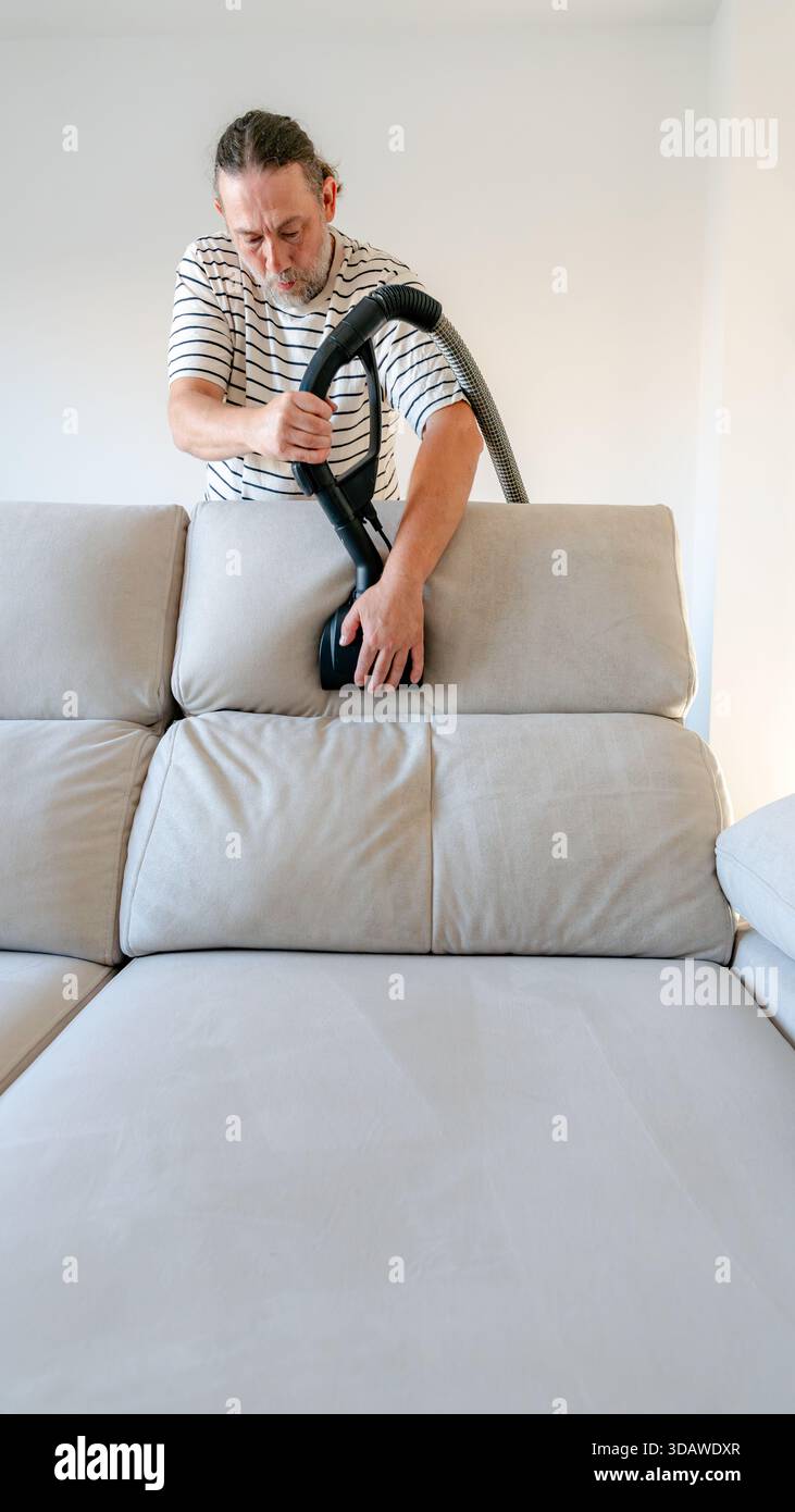 A mature man cleans his home sofa using a machine that utilizes pre-ionized water. This eco-friendly method avoids chemicals while ensuring the couch Stock Photo