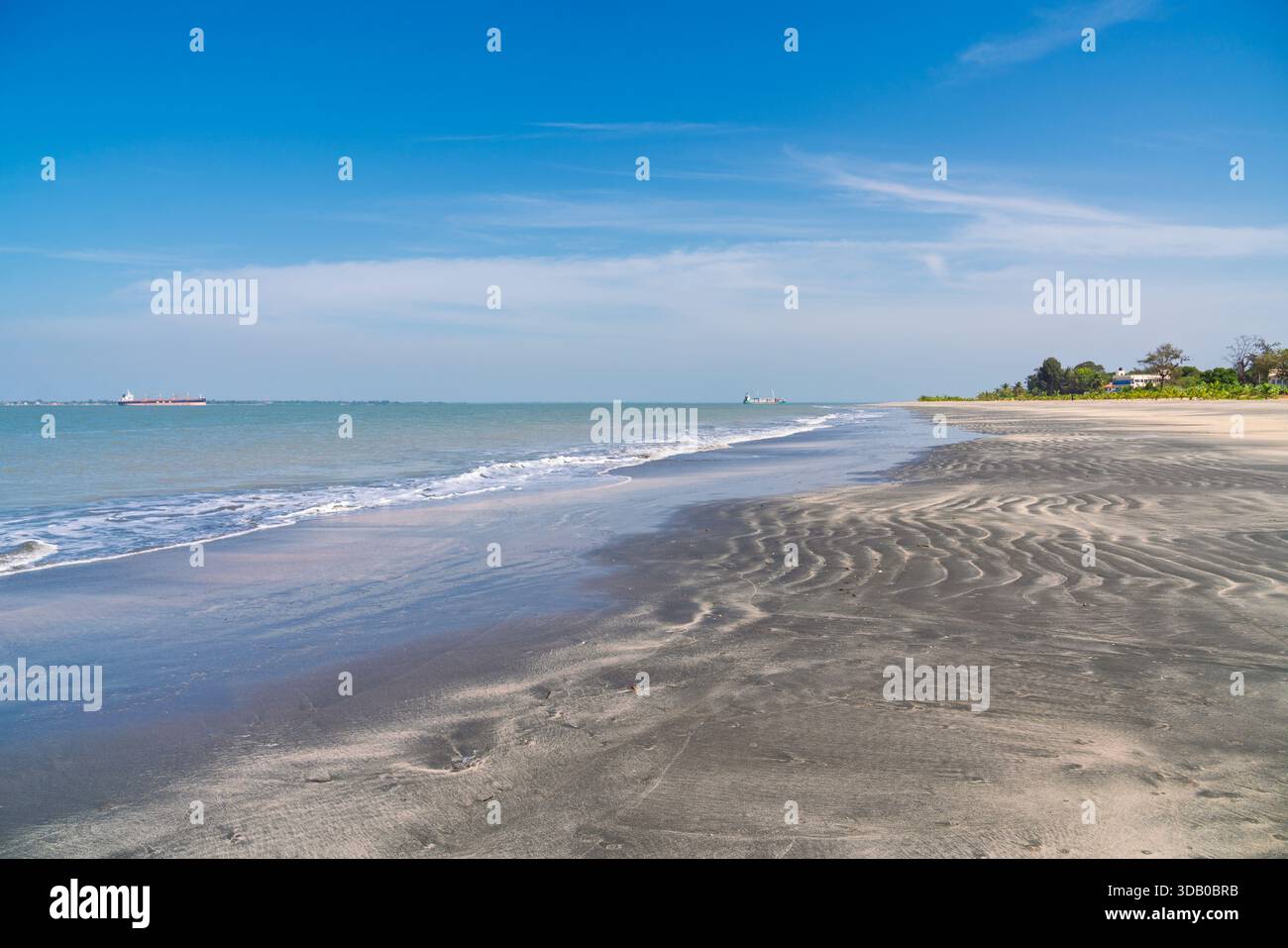 Wide, tranquil view of a tropical beach in Banjul, Gambia, featuring dark, rippled sand, small breaking waves, and clear blue sky Stock Photo