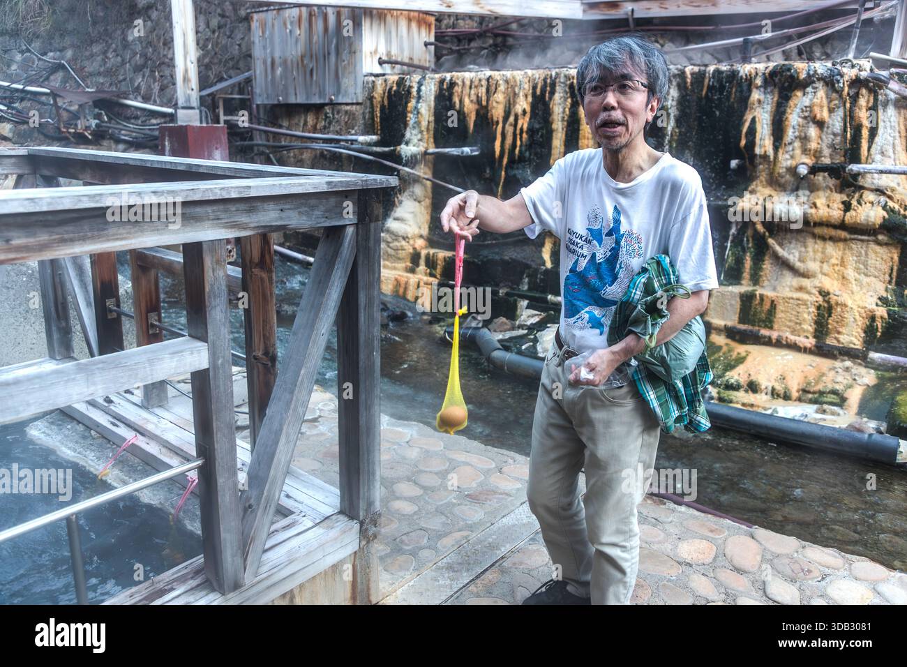 A tourist lowers raw eggs in a plastic net into the roughly 90°C spring water of the Yuzutsu in Yunomine Onsen, Tanabe, Wakayama Prefecture, Japan. On the riverbank, the water simmers in stone pools where eggs and vegetables are cooked in baskets or nets. Stock Photo
