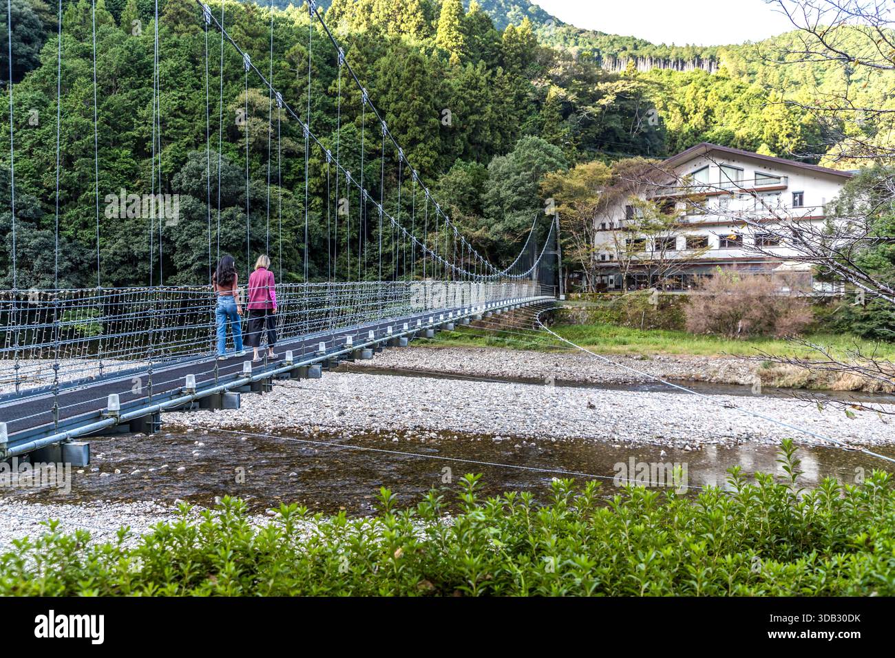 A suspension bridge crosses the Yomura River to the Watarase Onsen Hotel Sasayuri in the Hongu Valley. Two women walk across the bridge. Hongucho Wataze, Tanabe, Wakayama Prefecture, Japan. Stock Photo