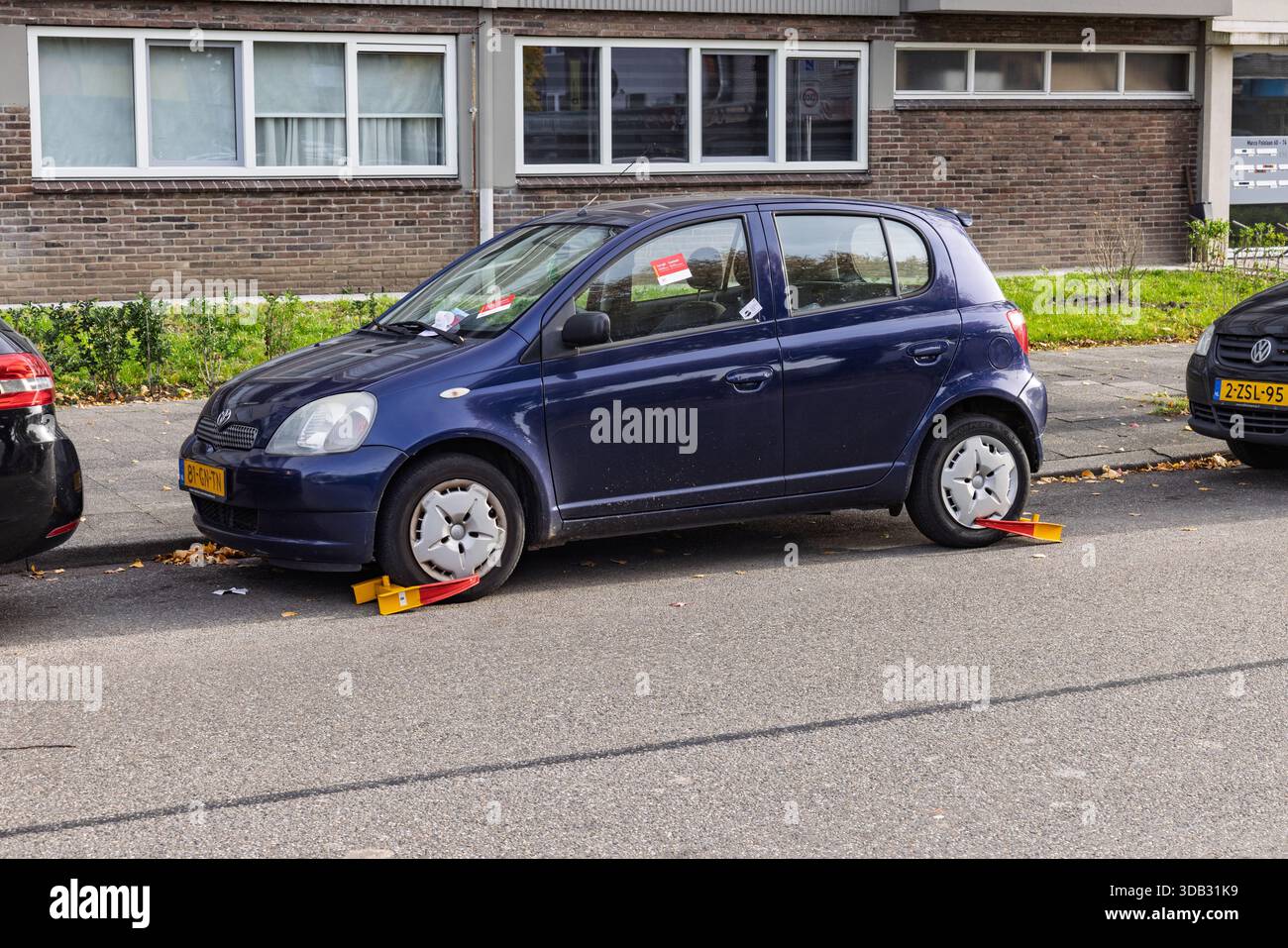 A parked blue car immobilized with two yellow and red wheel clamps on the tires due to a parking violation in a Dutch urban street setting. Stock Photo
