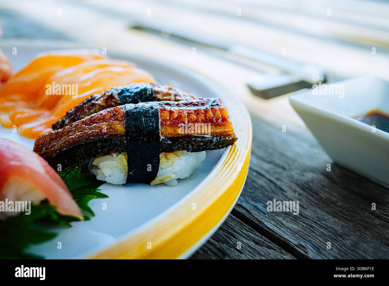 Unagi Nigiri sushi, featuring cooked eel glazed with sauce, rests alongside  salmon nigiri on a ceramic plate accented by a yellow rim. Wooden chopstic  Stock Photo - Alamy, image size:1300x956