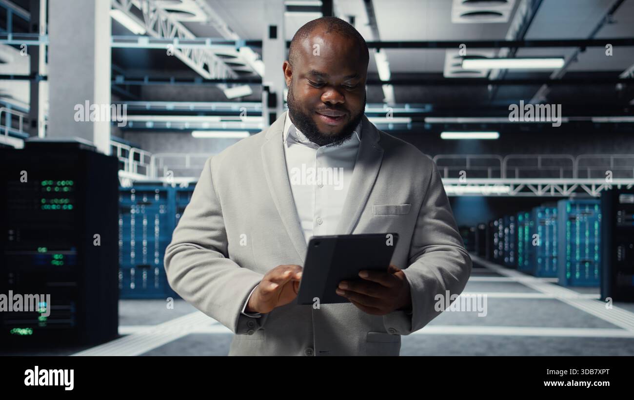 Smiling admin using tablet in in data center maintaining infrastructure for workloads. Joyful IT professional in server farm utilizing high tech server clusters for parallel processing, camera A Stock Photo