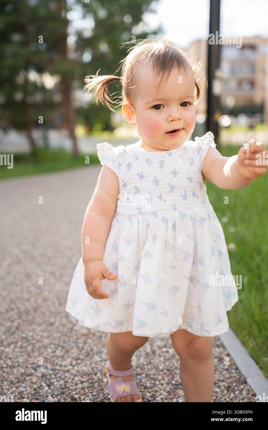 adorable toddler in white dress with blue accents walking on pebble path holding tiny flower, surrounded by greenery and blurred urban background on b Stock Photo - Alamy adorable toddler in white dress with blue accents walking on pebble path holding tiny flower, surrounded by greenery and blurred urban background on b Stock Photo - Alamy