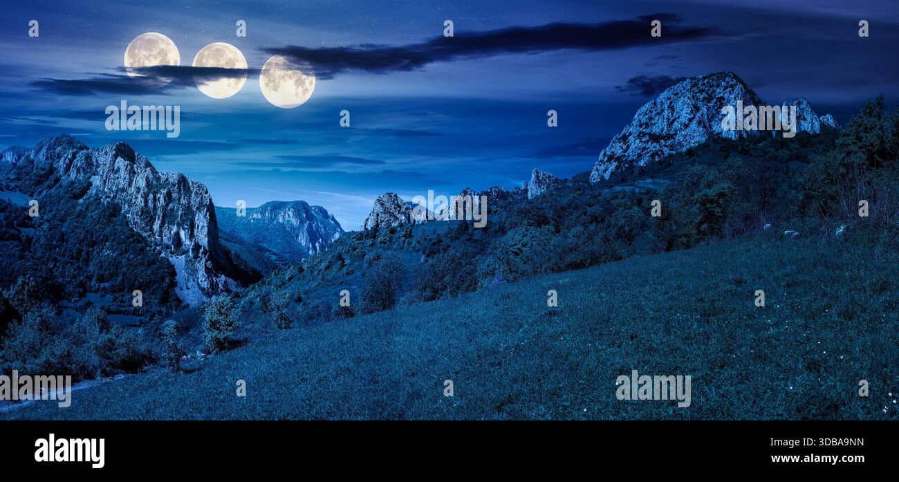 beautiful panorama of countryside in wonderful mountains at night. grassy meadow on hillside and distant gorge with hanging cliff in full moon light. Stock Photo