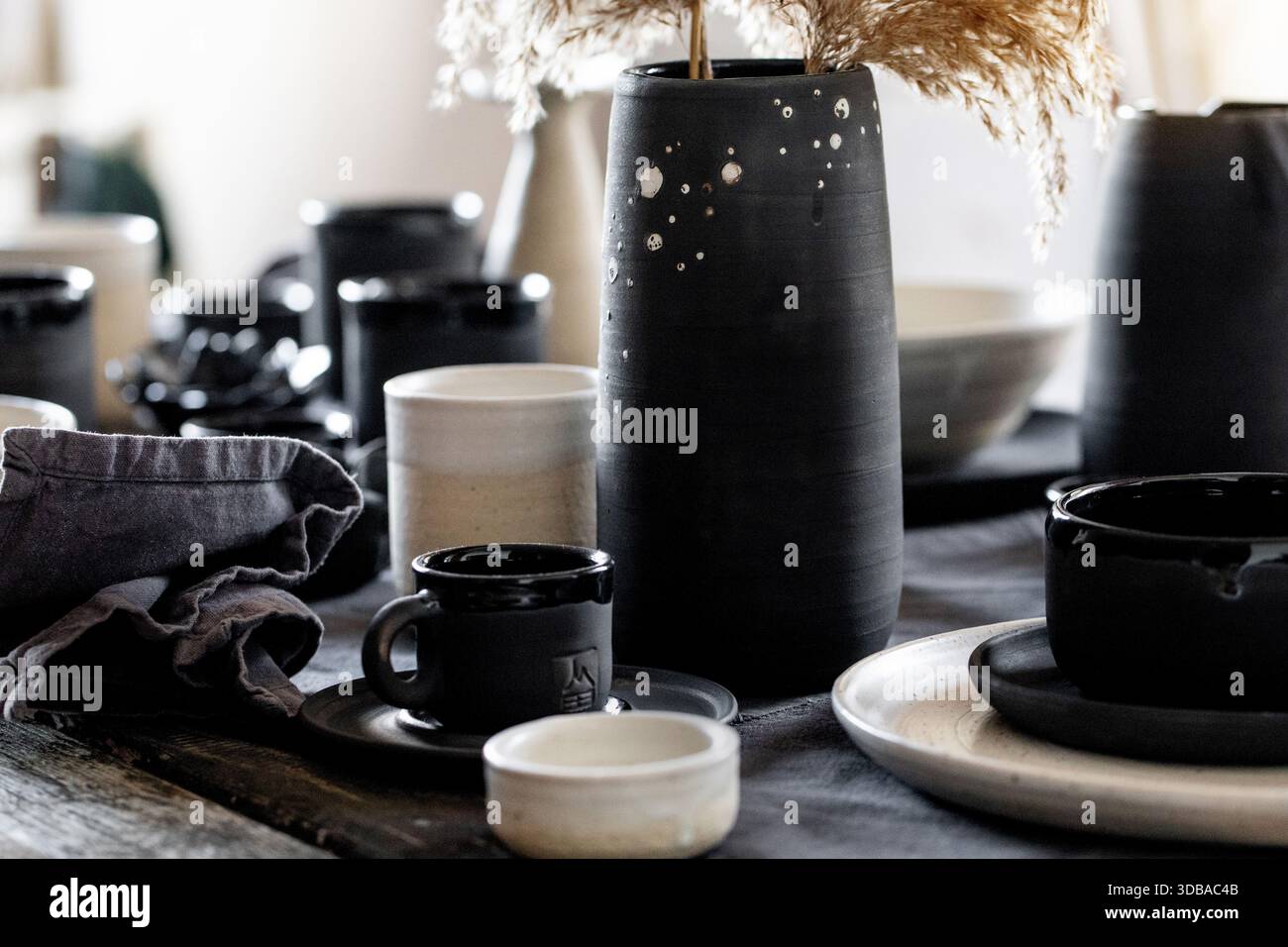 Rustic table setting with empty craft handmade ceramic tableware, black and white rough bowls, cups, jug and vase on linen tablecloth. Dry reed flower Stock Photo