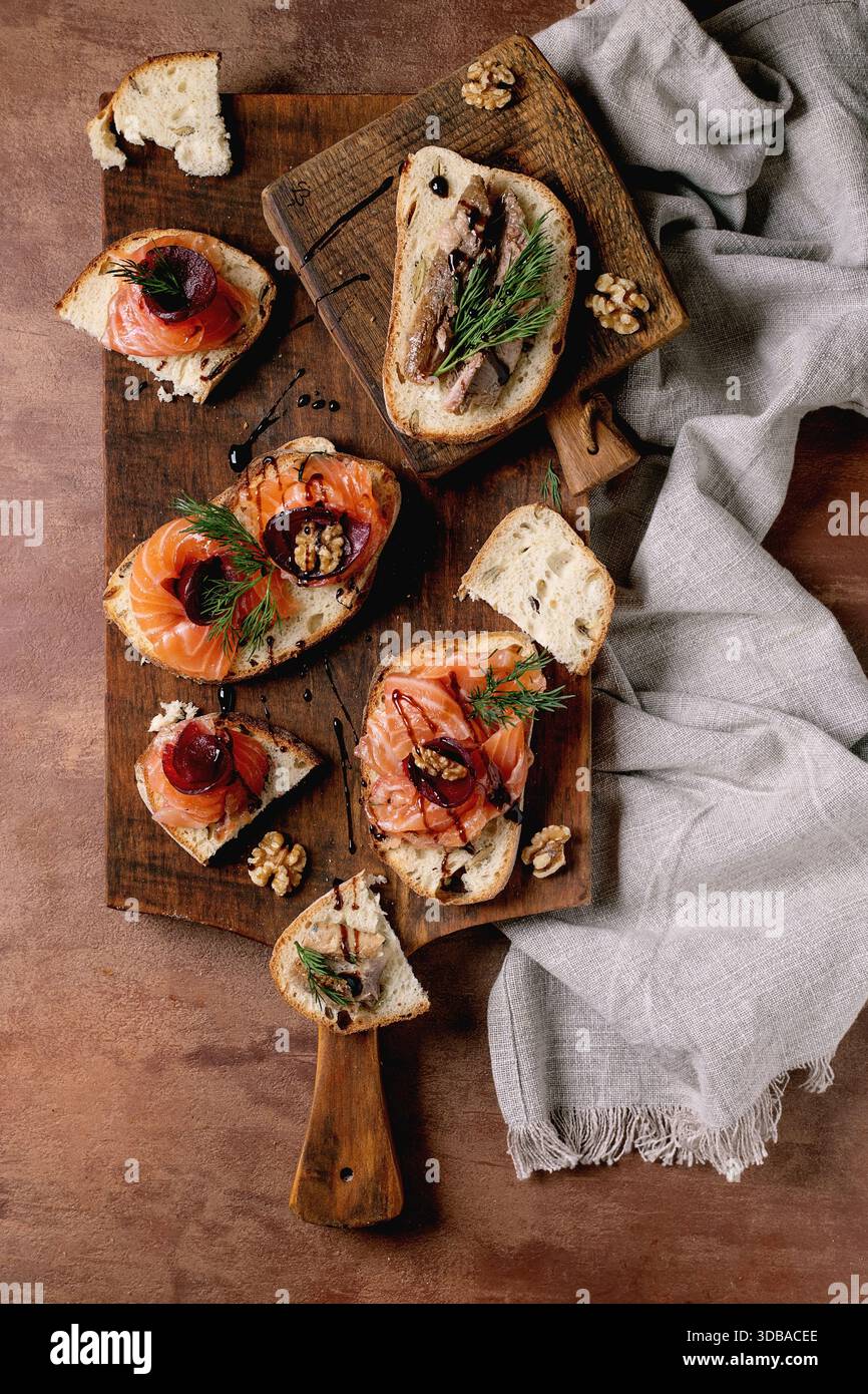 Sandwiches with thinly sliced salted salmon fillet, sardines, beetroot slices, walnuts, balsamic vinegar and fresh wheat bread on wooden board. Brown Stock Photo