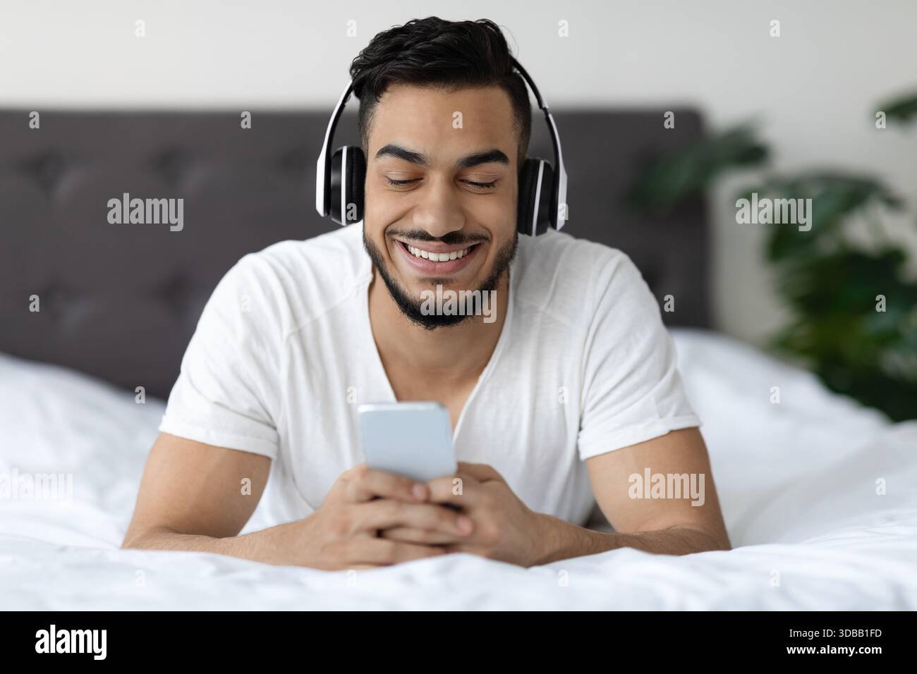 Happy Young Man Enjoys Technology While Relaxing in Bed With Smartphone Stock Photo