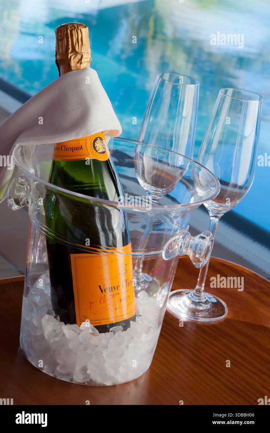 Veuve Clicquot fine vintage Champagne bottle with glasses, on ice in wine cooler, luxury sunlit infinity pool behind, with palm trees reflecting Stock Photo