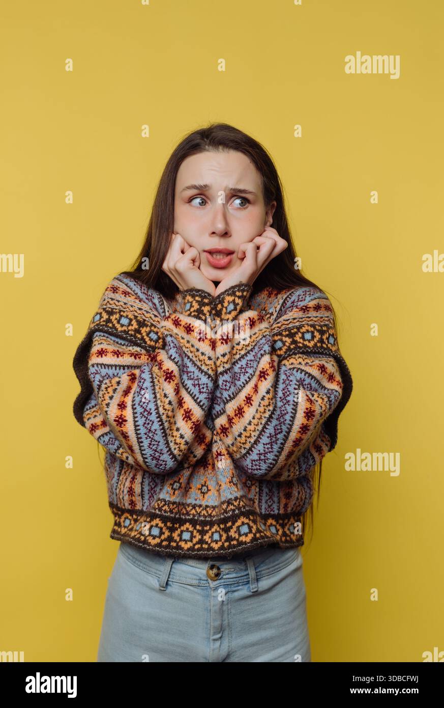 A woman stands against a bright yellow wall. She looks worried with her hands on her face. She wears a colorful patterned sweater and denim jeans. Her Stock Photo